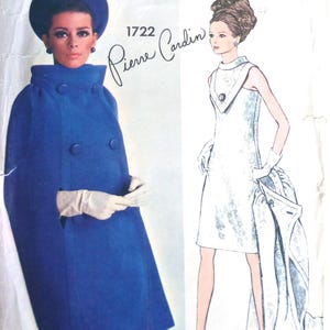 May include: A Vogue Paris Original sewing pattern envelope 1722 by Pierre Cardin. The front features a photograph of a person wearing a bright blue cape and matching hat, alongside a fashion illustration of a white sleeveless dress and coat.