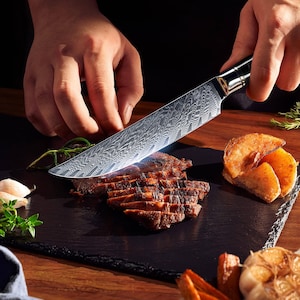 May include: A patterned steel chef's knife slicing grilled steak on a black slate board. The knife has a decorative handle and is cutting through the cooked meat. Roasted potatoes and herbs are also on the board, suggesting a culinary theme.