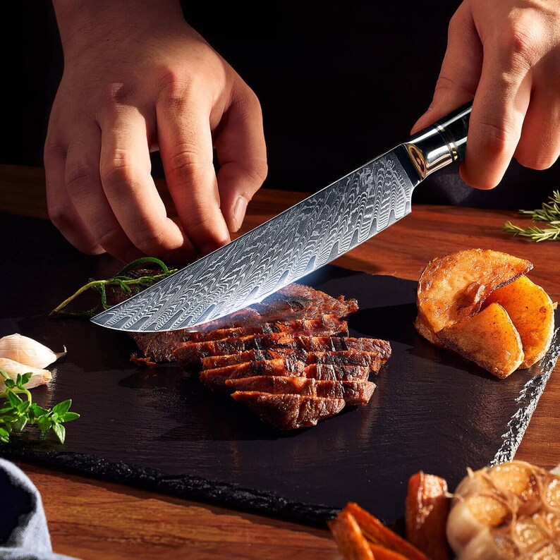 May include: A patterned steel chef's knife slicing grilled steak on a black slate board. The knife has a decorative handle and is cutting through the cooked meat. Roasted potatoes and herbs are also on the board, suggesting a culinary theme.