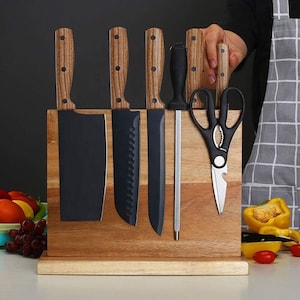 May include: A wooden magnetic knife holder with a stand, holding a set of black kitchen knives and a pair of kitchen shears. The holder is on a white countertop with fruit and vegetables.