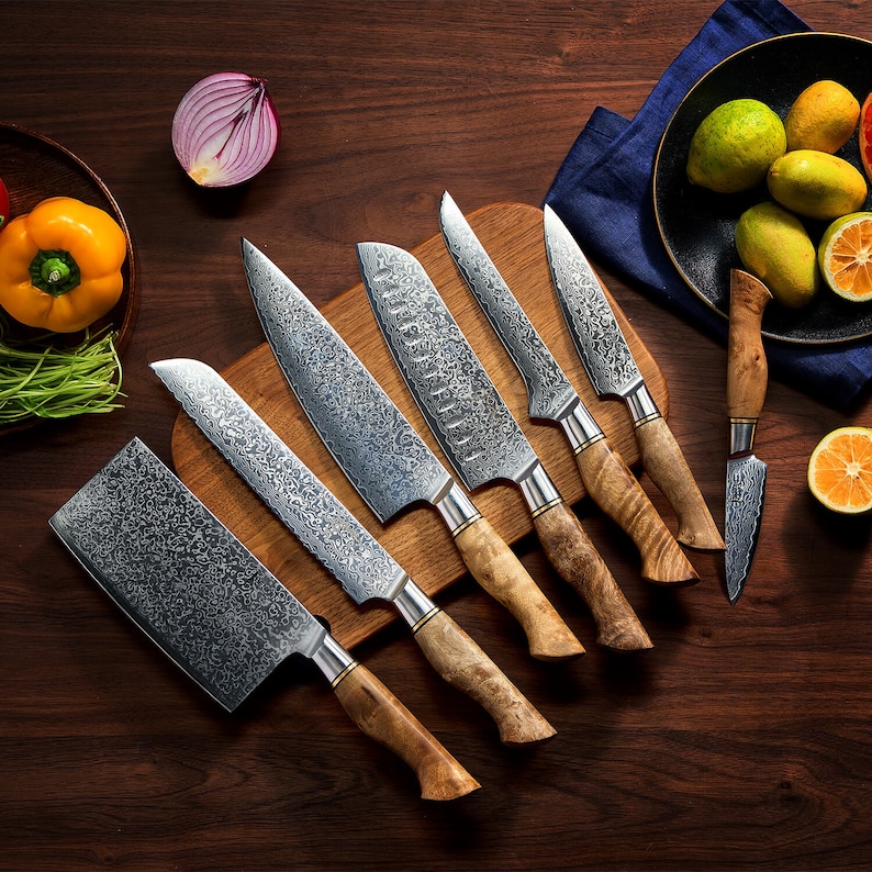 May include: A set of six kitchen knives with patterned Damascus steel blades and light brown wooden handles. The collection includes a cleaver, a chef's knife, a santoku knife, two utility knives, and a paring knife arranged on a wooden board.