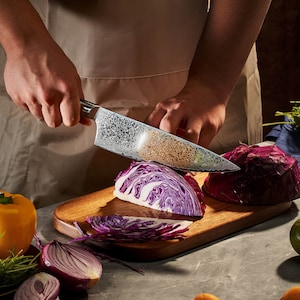 May include: A stainless steel chef knife with a patterned Damascus blade finish, being used to slice a head of red cabbage on a wooden cutting board, alongside a yellow bell pepper and a halved red onion on a gray surface.