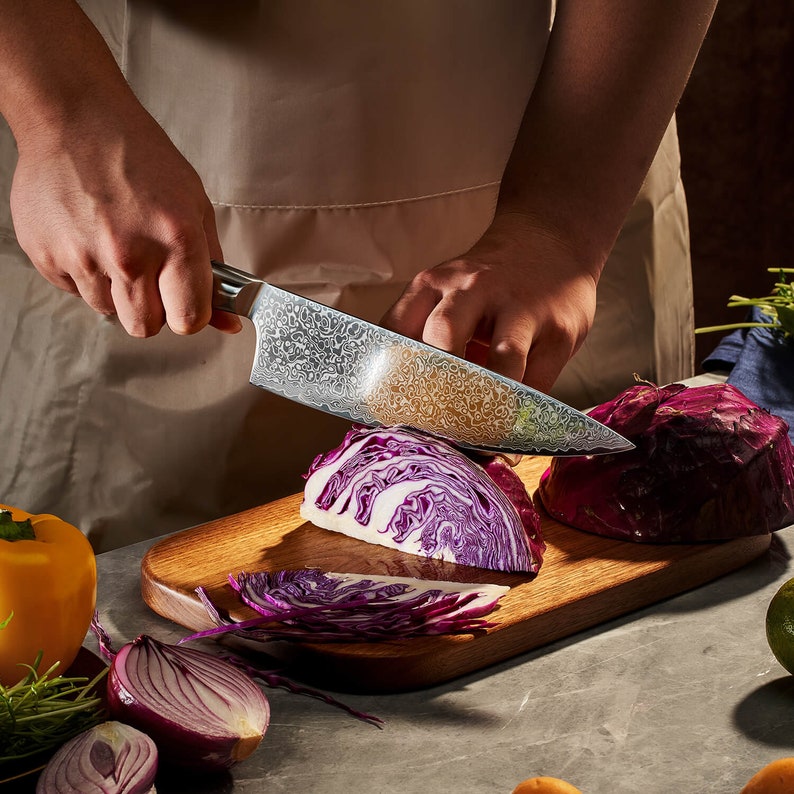 May include: A stainless steel chef knife with a patterned Damascus blade finish, being used to slice a head of red cabbage on a wooden cutting board, alongside a yellow bell pepper and a halved red onion on a gray surface.