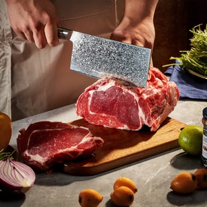 May include: A rectangular meat cleaver with a patterned Damascus steel blade is being used to slice a large piece of raw beef on a wooden cutting board. The scene includes a red onion, a lime, and small yellow fruits on the counter.