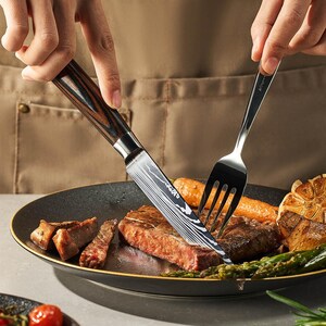 Pu&ograve; includere: Primo piano di una bistecca che viene tagliata con un coltello dal manico nero e marrone. La bistecca &egrave; su un piatto nero con asparagi e spicchi d'aglio.