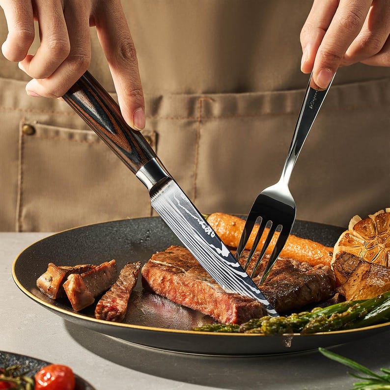 Pu&ograve; includere: Primo piano di una bistecca che viene tagliata con un coltello dal manico nero e marrone. La bistecca &egrave; su un piatto nero con asparagi e spicchi d'aglio.
