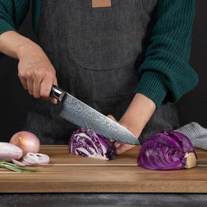 May include: A large, patterned chef's knife slicing through a head of purple cabbage on a wooden cutting board. Other vegetables, including red onions, are also present. The knife has a black handle.