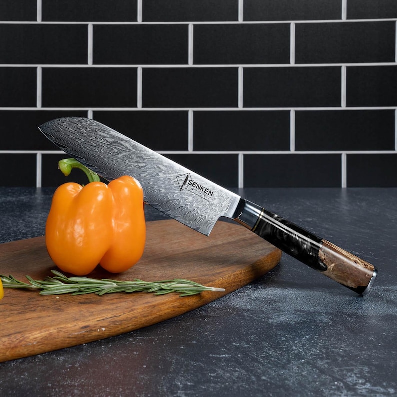 May include: A stainless steel chef's knife with a patterned blade and a dark wood handle rests on a wooden cutting board. An orange bell pepper and sprig of rosemary are next to the knife. The knife has the text "SENKEN" on the blade.