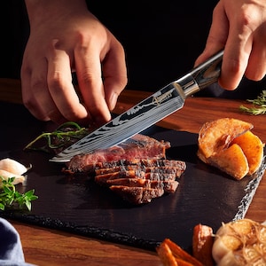 Pu&ograve; includere: Primo piano di una mano che tiene un coltello da cucina nero e argento con manico in legno, che taglia un pezzo di carne alla griglia su un tagliere di ardesia nero. La carne &egrave; tagliata a strisce sottili e ci sono altri pezzi di cibo sul tagliere, tra cui patate e aglio.
