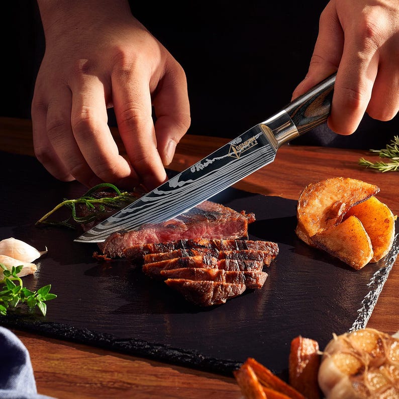 Pu&ograve; includere: Primo piano di una mano che tiene un coltello da cucina nero e argento con manico in legno, che taglia un pezzo di carne alla griglia su un tagliere di ardesia nero. La carne &egrave; tagliata a strisce sottili e ci sono altri pezzi di cibo sul tagliere, tra cui patate e aglio.