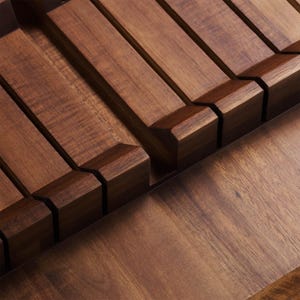 May include: A close-up shot of a wooden knife block. The block is made of dark brown wood and features slots for knives. The slots are angled and designed to hold knives securely. The wood has a smooth finish. The design is modern and minimalist.