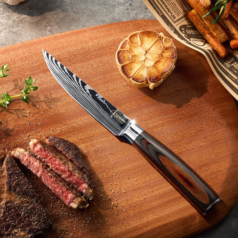 Pu&ograve; includere: Un primo piano di un coltello da cucina nero e argento con manico in legno. Il coltello &egrave; su un tagliere di legno con un pezzo di carne e uno spicchio d'aglio arrostito.