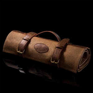 Puede incluir: Un rollo de cuchillos de lona marrón enrollado, con correas y asa de cuero. El rollo se asegura con dos correas de cuero y hebillas doradas. Un parche de cuero con el texto "SEIKEN" es visible en la parte delantera.