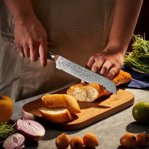 May include: A serrated bread knife with a patterned Damascus steel blade and a dark handle, slicing through a crusty loaf of bread on a wooden cutting board. The blade features a distinct wavy metal texture and a sharp, jagged edge.
