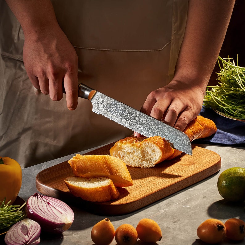 May include: A serrated bread knife with a patterned Damascus steel blade and a dark handle, slicing through a crusty loaf of bread on a wooden cutting board. The blade features a distinct wavy metal texture and a sharp, jagged edge.