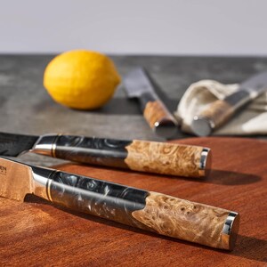 May include: Close-up of two kitchen knives with wooden handles and silver accents. The blades are stainless steel. A yellow lemon is in the background, along with two additional knives and a white cloth.