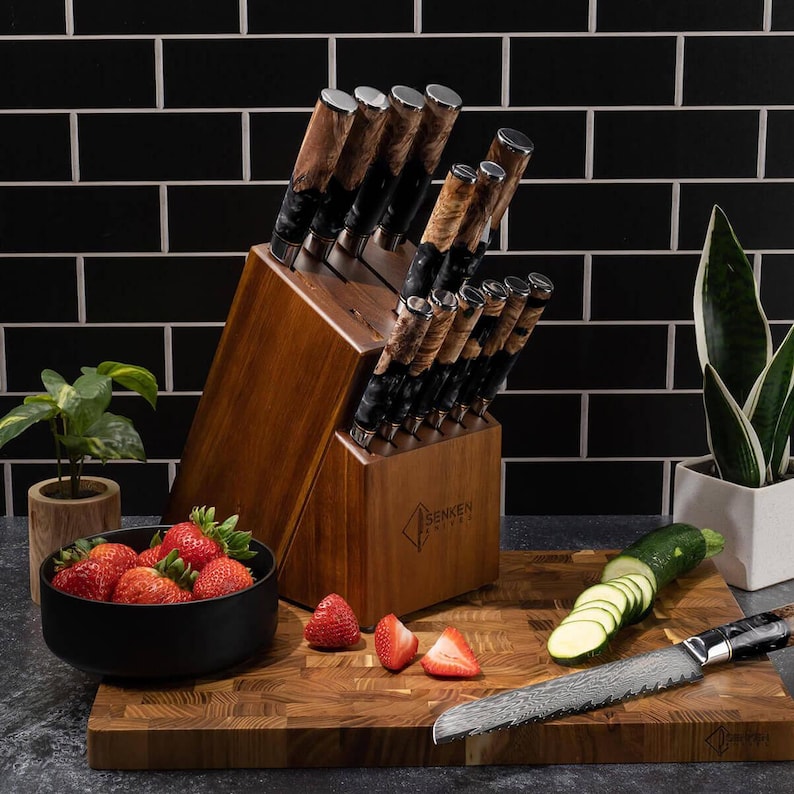 May include: A wooden knife block set with multiple knives, featuring brown and black handles. The set is displayed with a cutting board, strawberries, and sliced zucchini. The knives have a unique patterned blade design.