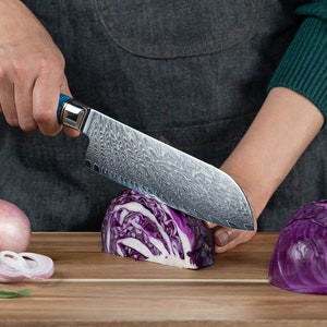 Pode incluir: Uma pessoa está cortando um repolho roxo com uma faca de cozinha prateada e azul em uma tábua de cortar de madeira. Também há algumas cebolas vermelhas na tábua.