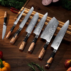 May include: A set of six kitchen knives with patterned steel blades and wooden handles. The knives are arranged on a wooden cutting board, showcasing their various sizes and blade styles. The handles feature a mix of black and light brown wood.