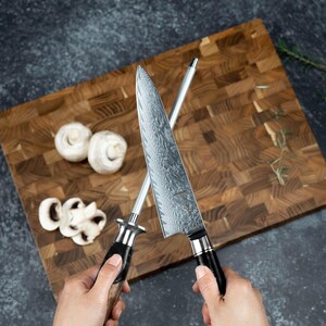 K&ouml;nnte beinhalten: Ein Kochmesser mit gemusterter Klinge und ein Wetzstahl werden &uuml;ber einem Holzschneidebrett mit Pilzen gehalten. Das Messer hat einen schwarzen Griff. Das Schneidebrett besteht aus Holzbl&ouml;cken.