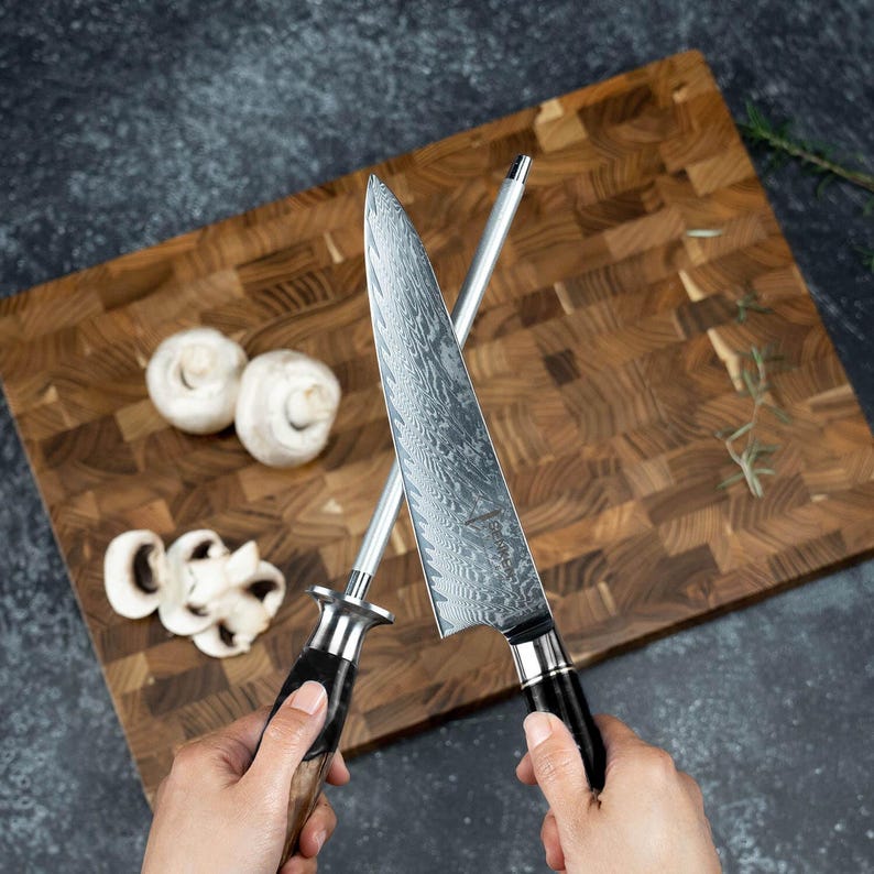 K&ouml;nnte beinhalten: Ein Kochmesser mit gemusterter Klinge und ein Wetzstahl werden &uuml;ber einem Holzschneidebrett mit Pilzen gehalten. Das Messer hat einen schwarzen Griff. Das Schneidebrett besteht aus Holzbl&ouml;cken.