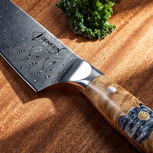 Puede incluir: Primer plano de un cuchillo de chef de acero inoxidable con una hoja estampada y un mango de madera. El cuchillo tiene la palabra "SENKEN" impresa en la hoja. El mango presenta un dise&ntilde;o decorativo con un acento plateado.