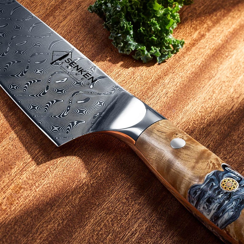 Puede incluir: Primer plano de un cuchillo de chef de acero inoxidable con una hoja estampada y un mango de madera. El cuchillo tiene la palabra "SENKEN" impresa en la hoja. El mango presenta un dise&ntilde;o decorativo con un acento plateado.