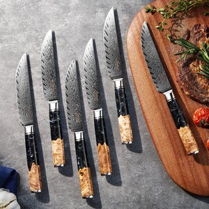 May include: A set of six steak knives with patterned steel blades and handles featuring a combination of black, white, and light brown wood. The knives are arranged on a gray surface, with one knife next to a wooden cutting board with food.