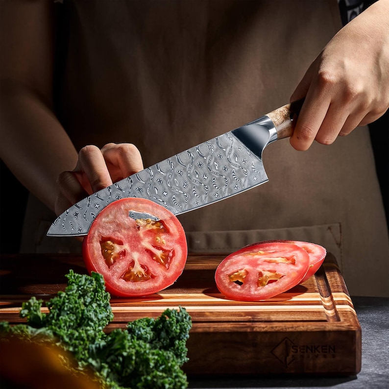 Puede incluir: Un cuchillo de chef de acero inoxidable con una hoja estampada corta un tomate rojo sobre una tabla de cortar de madera. El cuchillo tiene un mango de madera. Se ven rodajas de tomate. La col rizada verde est&aacute; en primer plano.