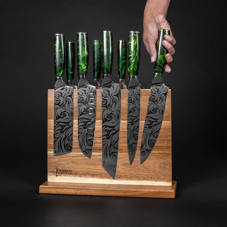 May include: A set of eight black and green kitchen knives with decorative etched designs on the blades. The knives are displayed in a wooden magnetic knife holder.