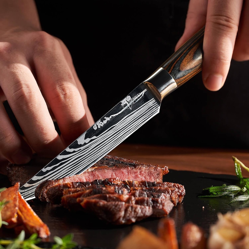 Pu&ograve; includere: Primo piano di una mano che tiene un coltello con manico in legno e argento con una lama a motivi, che taglia un pezzo di carne alla griglia.