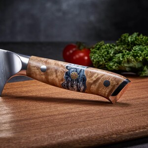 Puede incluir: Primer plano de un cuchillo de chef con mango de madera. El mango presenta una incrustaci&oacute;n decorativa con detalles azules y dorados. La hoja es de acero inoxidable. El cuchillo descansa sobre una tabla de cortar de madera, con un tomate y col rizada en el fondo.