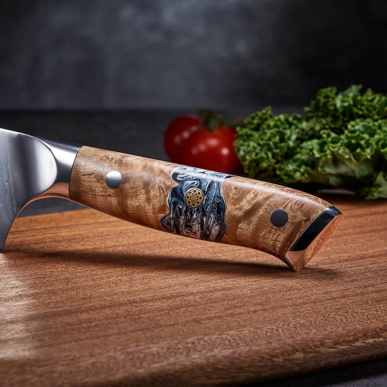 Puede incluir: Primer plano de un cuchillo de chef con mango de madera. El mango presenta una incrustaci&oacute;n decorativa con detalles azules y dorados. La hoja es de acero inoxidable. El cuchillo descansa sobre una tabla de cortar de madera, con un tomate y col rizada en el fondo.