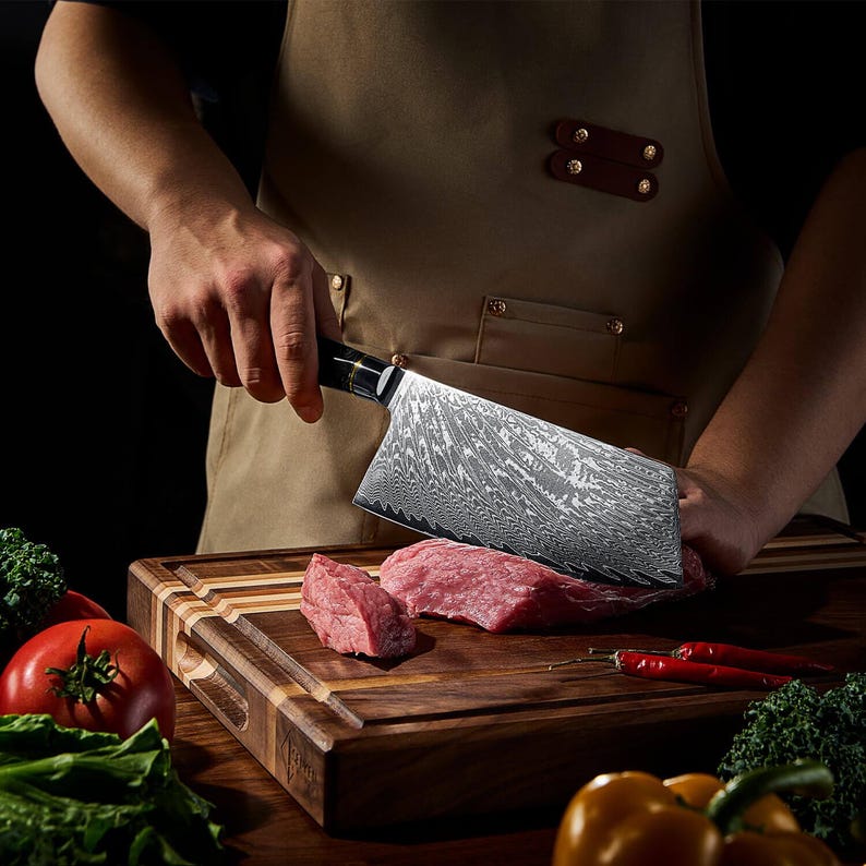 May include: A large, patterned cleaver knife cutting raw meat on a wooden cutting board. The knife has a black handle and a silver blade. Red chili peppers, tomatoes, and broccoli are also visible.