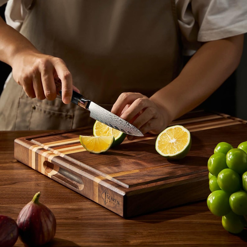 May include: A person slicing a lime on a wooden cutting board with a patterned knife. The cutting board has a variety of wood tones and a handle. The knife has a black handle and a patterned blade. Limes and green grapes are also visible.