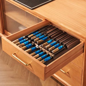 May include: A collection of steak knives with blue and woodgrain handles, presented in a wooden drawer. The handles feature a striking blue resin and woodgrain design. The drawer is part of a light-coloured wooden cabinet with a gold-toned handle.