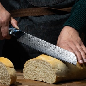 May include: A person is using a silver bread knife with a patterned blade to cut a loaf of white bread. The knife has a black handle with a blue and white pattern.