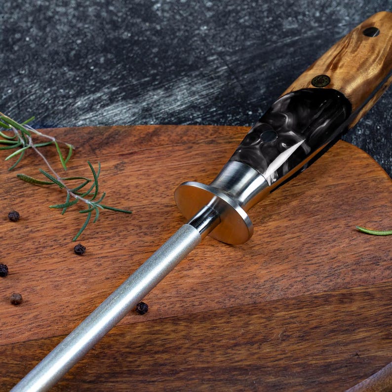 May include: A close-up of a knife sharpener with a wooden handle and a silver steel rod. The handle has a black and brown marbled design. The sharpener is on a wooden cutting board with rosemary and peppercorns.