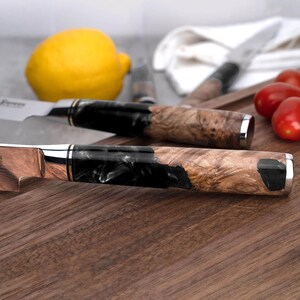May include: Close-up of two kitchen knives with wooden and black resin handles. The blades are silver and copper-toned, with a silver bolster. The handles have a natural wood grain pattern. A lemon and tomatoes are in the background.