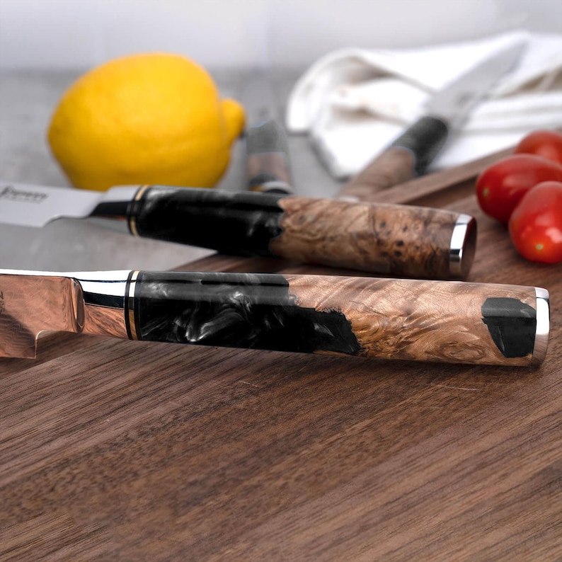 May include: Close-up of two kitchen knives with wooden and black resin handles. The blades are silver and copper-toned, with a silver bolster. The handles have a natural wood grain pattern. A lemon and tomatoes are in the background.