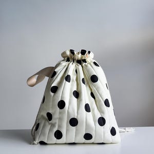 May include: A cream-coloured drawstring bag featuring a repeating pattern of black polka dots. The bag is constructed with vertical quilted stitching and includes a beige fabric handle on the side, with white cord strings used to close the top opening.