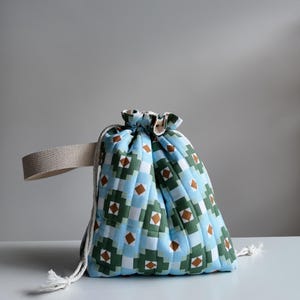 Può includere: Una pochette trapuntata con chiusura a coulisse caratterizzata da un motivo geometrico nei colori azzurro, verde scuro e marrone. La borsa presenta un manico in tessuto beige sul lato e cordini bianchi con estremità sfilacciate per la chiusura.