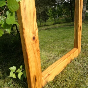 Op de afbeelding: Een grote, rechthoekige spiegel met een natuurlijke houten lijst. De lijst is gemaakt van dik, onbewerkt hout, dat de natuurlijke nerf en knopen laat zien. De spiegel staat buiten op een grasveld, met bomen en bladeren op de achtergrond.