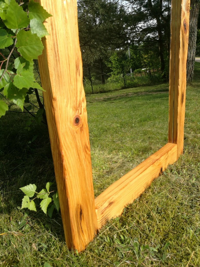 Op de afbeelding: Een grote, rechthoekige spiegel met een natuurlijke houten lijst. De lijst is gemaakt van dik, onbewerkt hout, dat de natuurlijke nerf en knopen laat zien. De spiegel staat buiten op een grasveld, met bomen en bladeren op de achtergrond.