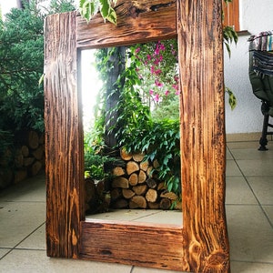 Puede incluir: Un espejo de madera r&uacute;stico con un marco rectangular. El marco est&aacute; hecho de madera recuperada con un acabado marr&oacute;n oscuro. El espejo refleja una vista de follaje verde y una pila de troncos.