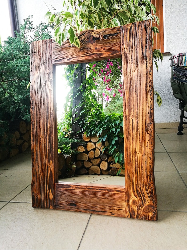Puede incluir: Un espejo de madera r&uacute;stico con un marco rectangular. El marco est&aacute; hecho de madera recuperada con un acabado marr&oacute;n oscuro. El espejo refleja una vista de follaje verde y una pila de troncos.