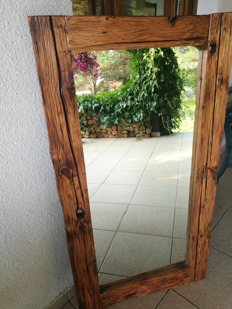 Puede incluir: Un espejo de madera r&uacute;stico con un marco rectangular. El marco est&aacute; hecho de madera recuperada con un acabado marr&oacute;n natural. El espejo refleja una vista de un patio con plantas verdes y un suelo de piedra.