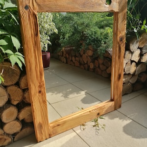Op de afbeelding: Een groot, rechthoekig houten frame met een natuurlijke, lichtbruine afwerking. Het frame is gemaakt van dik, getextureerd hout, met zichtbare nerven en knopen. Het frame leunt tegen een betegeld oppervlak, met groen op de achtergrond.