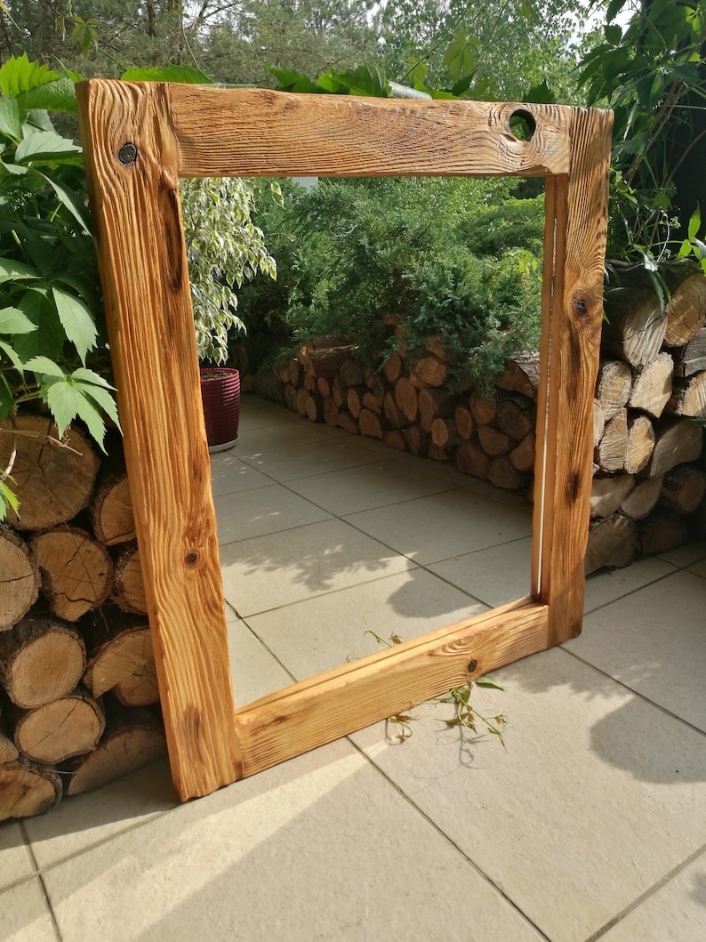 Op de afbeelding: Een groot, rechthoekig houten frame met een natuurlijke, lichtbruine afwerking. Het frame is gemaakt van dik, getextureerd hout, met zichtbare nerven en knopen. Het frame leunt tegen een betegeld oppervlak, met groen op de achtergrond.