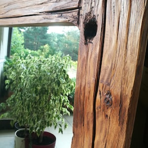 Puede incluir: Un marco de espejo de madera r&uacute;stico con un acabado de madera natural. El marco tiene un nudo y una grieta en la madera, lo que aumenta su car&aacute;cter. El espejo refleja una planta verde en una maceta.
